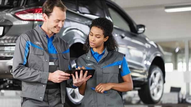 Two auto technicians reviewing information on a tablet in a service workshop with a car lifted in the background.