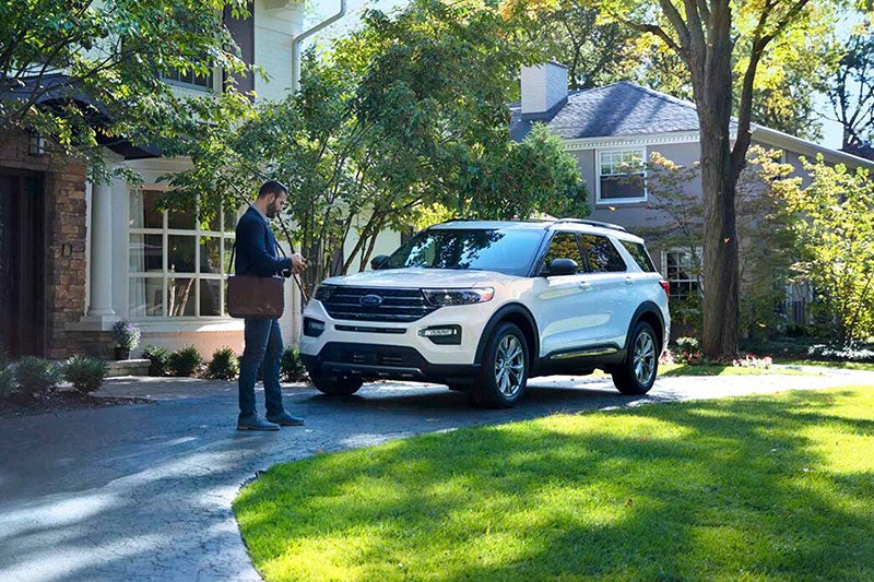 A man with a satchel stands by a white Ford Explorer SUV in a driveway, looking at his phone.