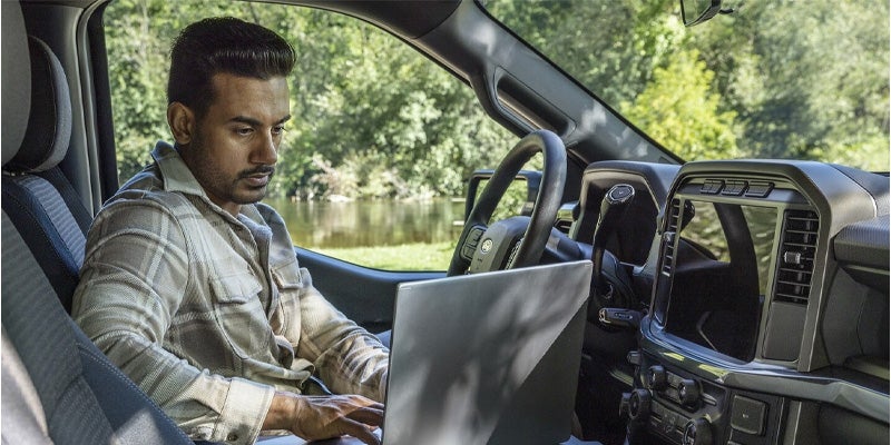 A man sits inside a vehicle, typing on a laptop with trees visible outside the window.
