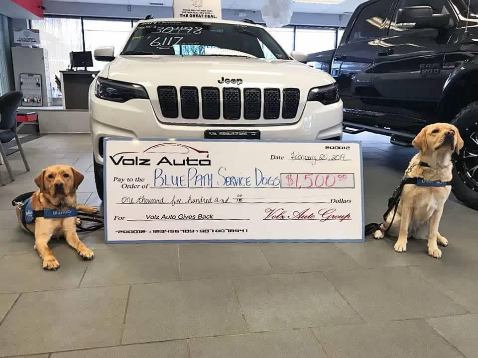Two service dogs sitting beside an oversized donation check in front of a white Jeep.