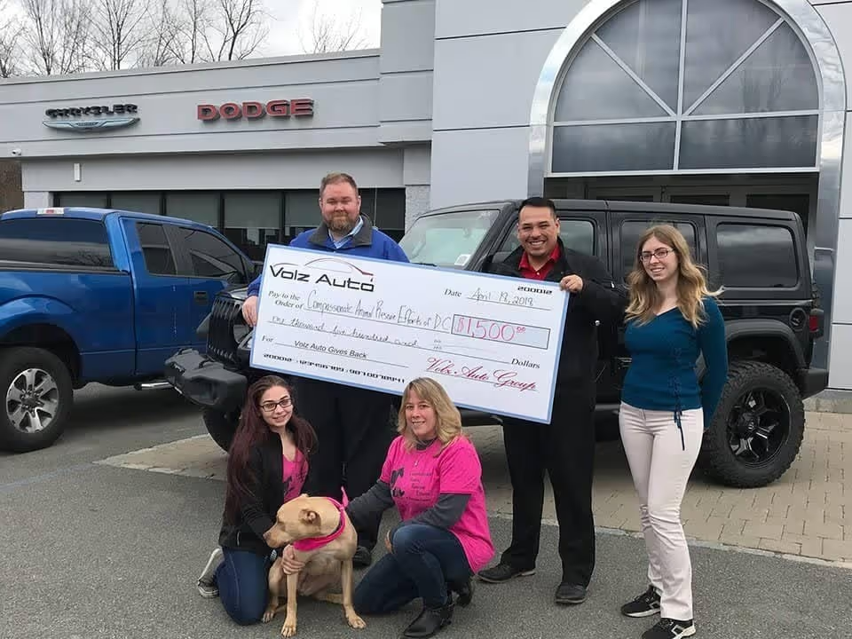 People and a dog posing outside a dealership with a donation check