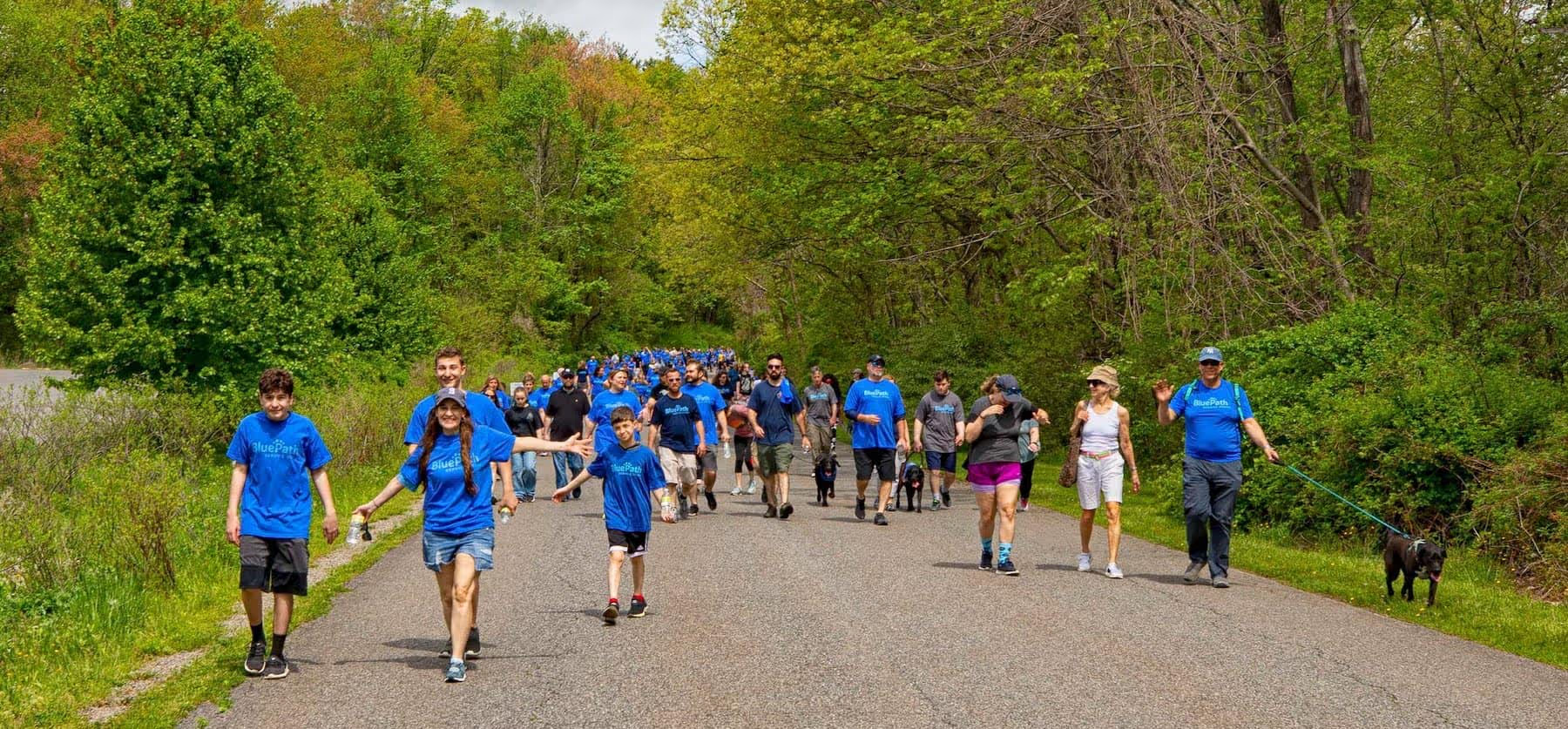 A long line of about 50 people and a few dogs walk away from the viewer on a paved road surrounded by green trees.
