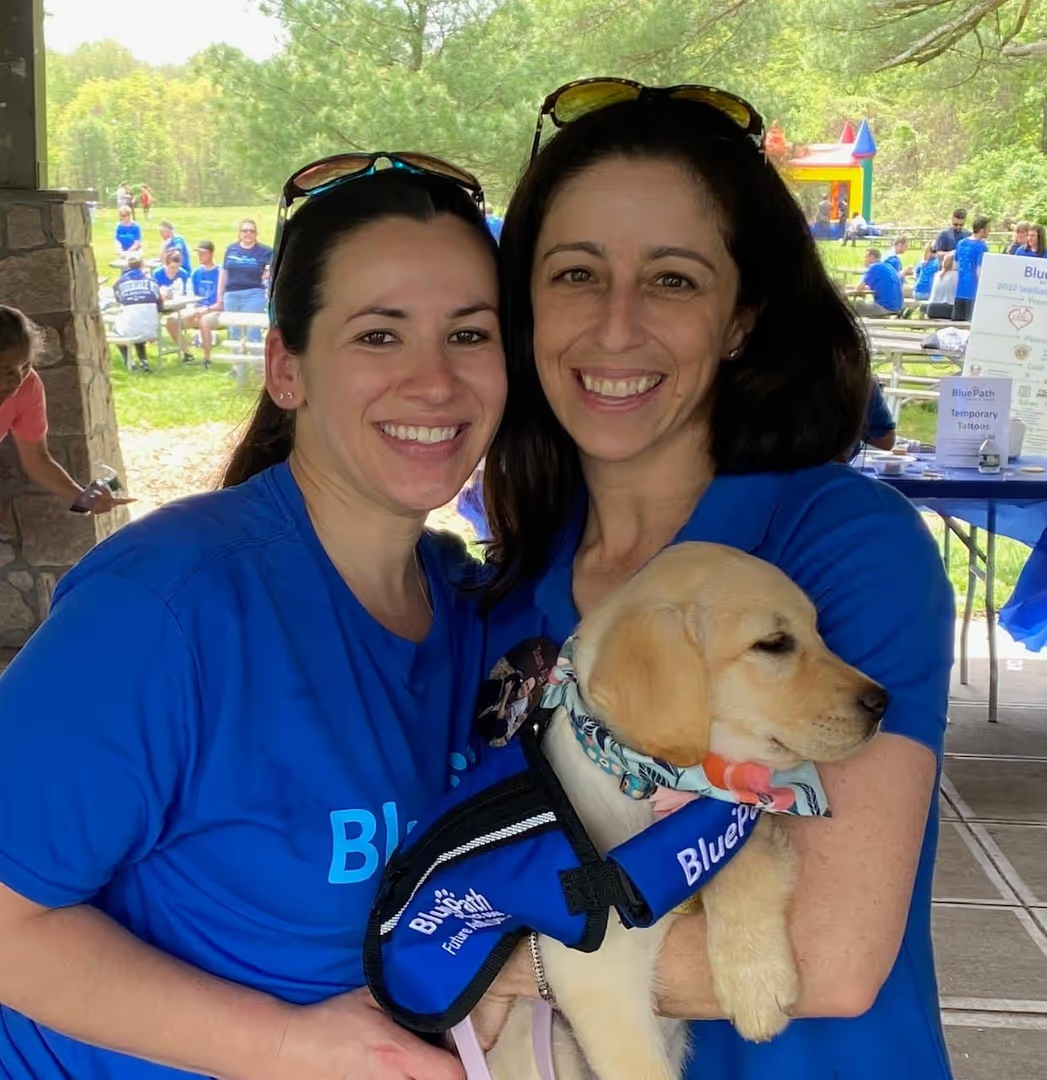Two smiling women in blue 'Blue Path' shirts hold a blonde Labrador puppy in a blue training vest. Green park background.